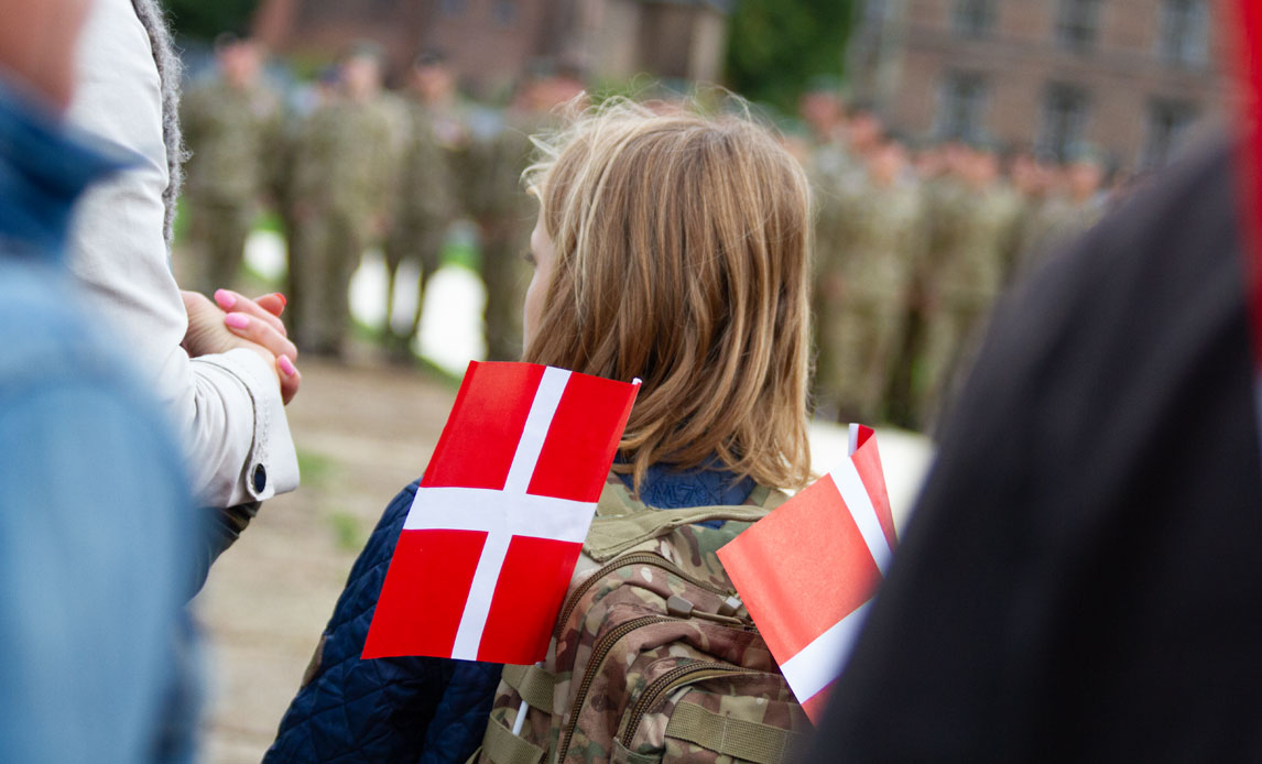 En pige står med ryggen til kameraet med en militær rygsæk på, hvor der er to dannebrogsflag i. I forgrunden kan man se soldater i parade.
