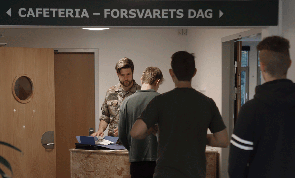 Tre unge mænd står i kø til at få registreret deres fremmøde til Forsvarets Dag/session.
En officer i uniform står for registreringen