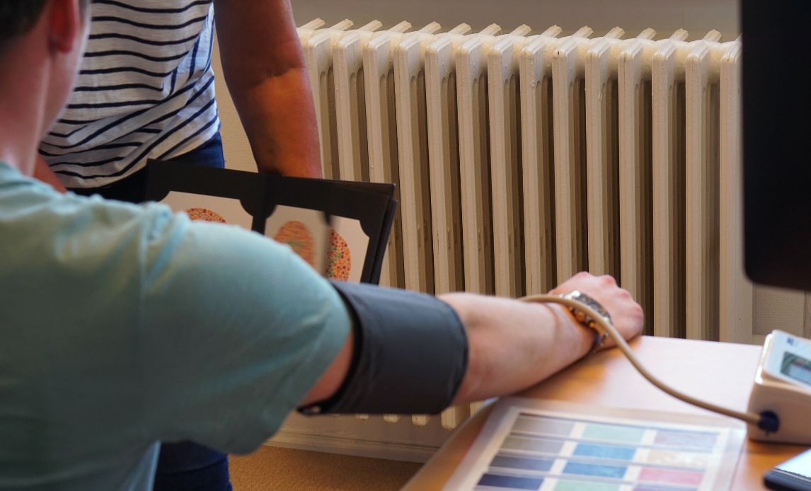 På billedet ses armen af en ansøger til HRU, der får målt blodtryk, mens en kvindelig medarbejder viser ham en række farvede tal for at teste, om ansøgeren er farveblind.