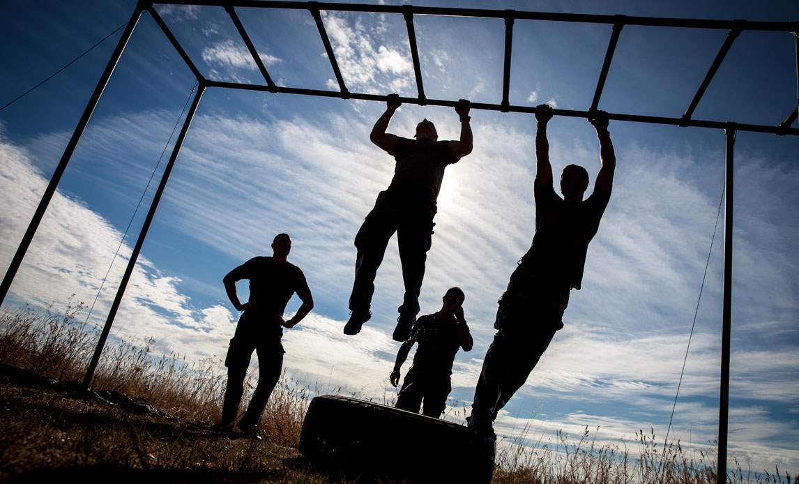 Fire elever fra Frømandskorpsets elevskole træner arme på en vandret stige. Billedet er taget i frøperspektiv og i modlys, så man ikke kan se deres ansigter.
