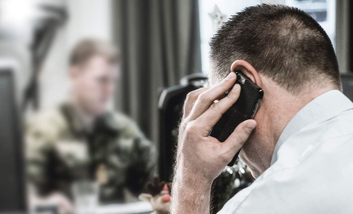 En mandlig medarbejder med ryggen til taler i telefon. I baggrunden kan se omridset af en soldat foran en computer