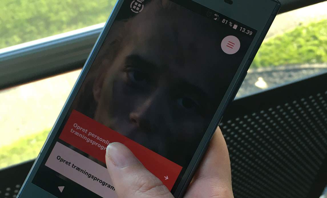 Nærbillede af en hånd der holder en mobiltelefon, hvor man se forsiden af appen Træn med Forsvaret