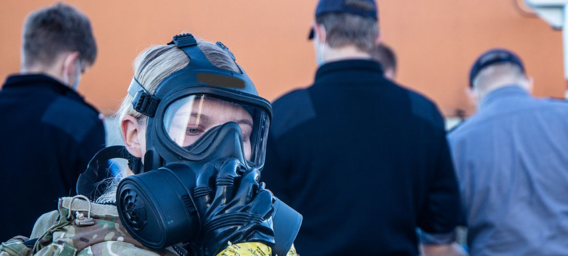 I baggrunden er en orange lastbil, forrest står en lyshåret kvinde i uniform med en røgmaske.
Bag hende står fire kolleger med ryggen til.