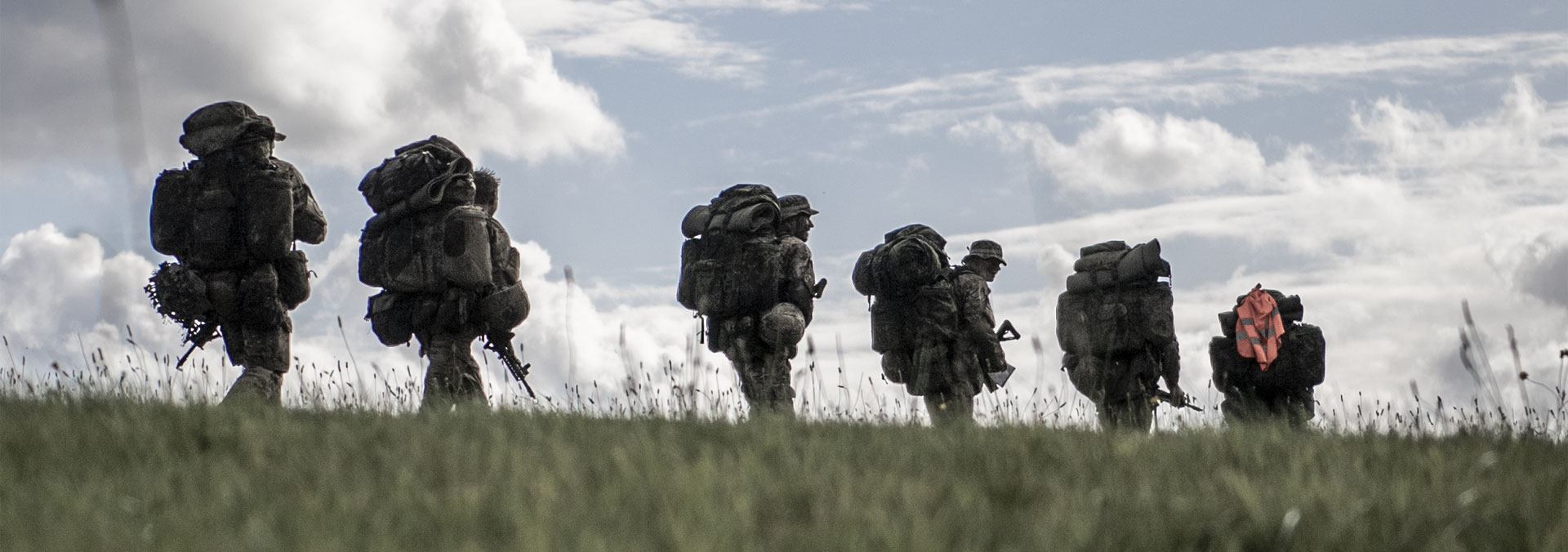 Soldater med oppakning marcherer over en mark. Billedet er taget med afstand, lidt i frøperspektiv og der er grønt græs i forgrunden.
