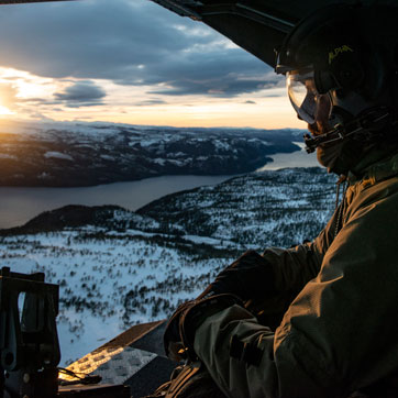En soldat med hjelm og headset sidder i en helikopter og kigger udover et snebeklædt landskab