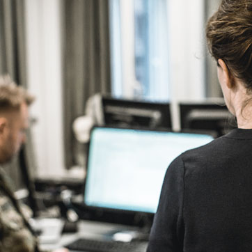 En kvindelig medarbejder står med ryggen til kameraet og kigger på en mandlig kollega i uniform, der sidder for sin computer