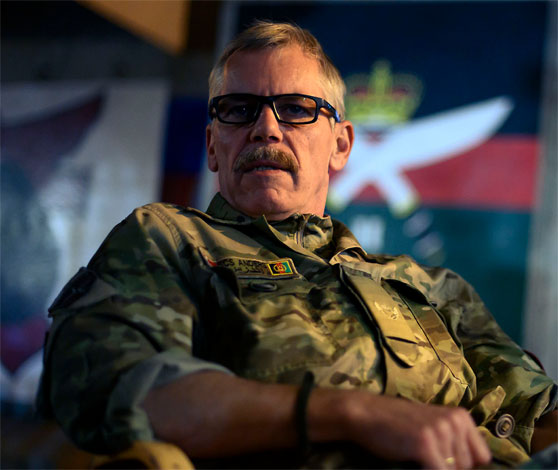 En mand i uniform sidder i en stol med flag i baggrunden