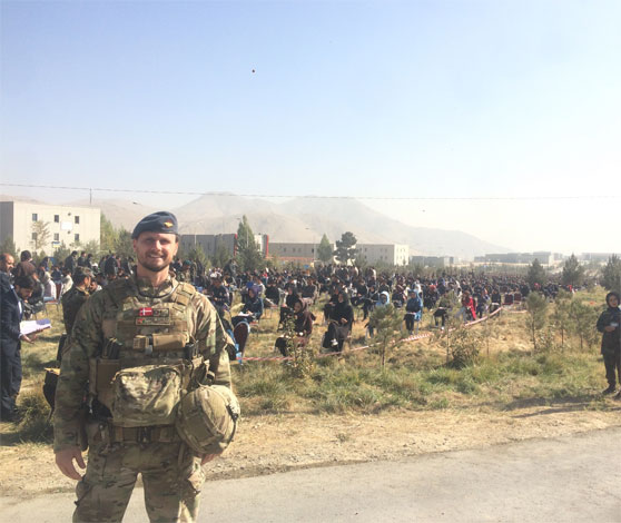 En smilende soldat fotograferet i Afghanistan med lokalbefolkning, bjerge og blå himmel i baggrunden